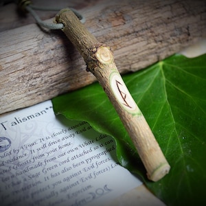 May include: A wooden talisman with a green cord and a carved rune symbol. The talisman is on a piece of driftwood and a green leaf. The text on the paper reads "c Wood Talisman".