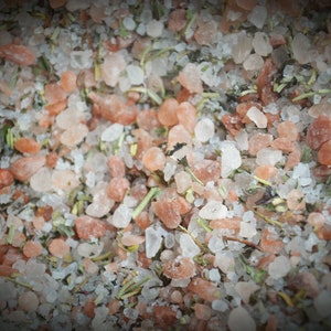 May include: Close-up of a mixture of pink Himalayan salt, white sea salt, and dried herbs. The salt crystals are various sizes and shapes, and the herbs are a mix of green and brown.
