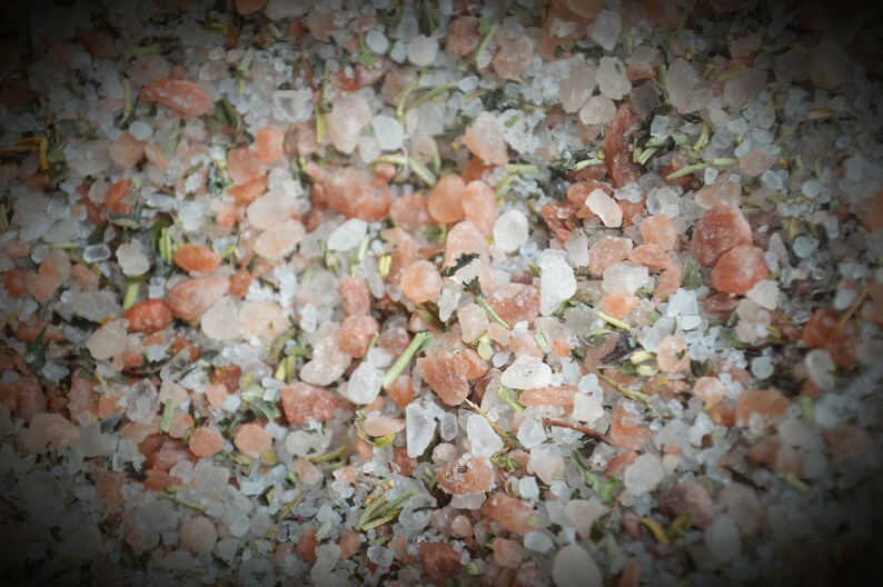 May include: Close-up of a mixture of pink Himalayan salt, white sea salt, and dried herbs. The salt crystals are various sizes and shapes, and the herbs are a mix of green and brown.