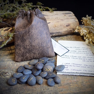May include: A set of blue rune stones with gold markings, a brown drawstring bag, and a handwritten note on a wooden surface. The bag has a textured appearance, and the stones are oval. Dried plants and wood are in the background.