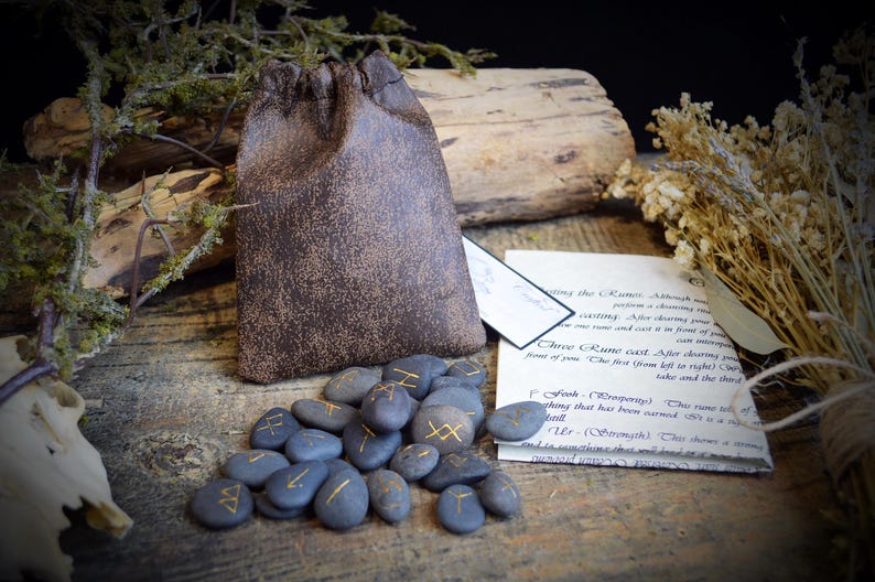 May include: A collection of blue rune stones with gold symbols, a brown drawstring bag, and a handwritten note on a wooden surface. The bag is textured, and the stones are oval-shaped. Dried plants and a piece of wood are in the background.