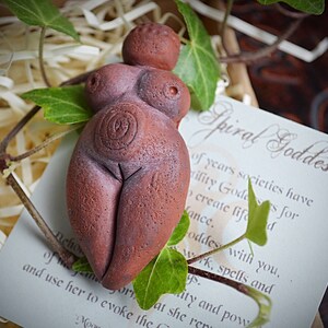 May include: A terracotta figurine of a spiral goddess, with a spiral design on her belly. The figurine is sitting on a white card with the text "Spiral Goddess" written in cursive.