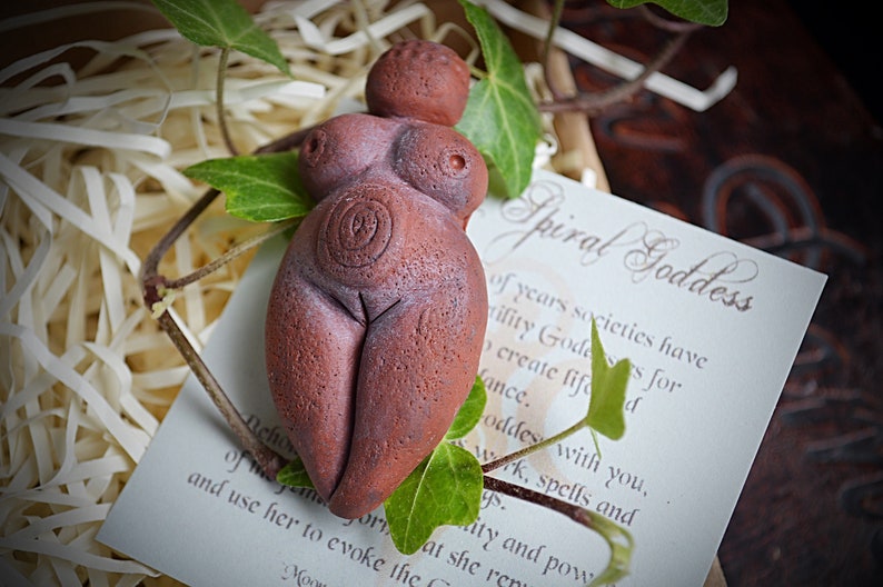May include: A terracotta figurine of a spiral goddess, with a spiral design on her belly. The figurine is sitting on a white card with the text "Spiral Goddess" written in cursive.
