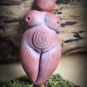 May include: A terracotta figurine of a female figure with a circular design on the abdomen. The figurine is standing on a bed of green moss.