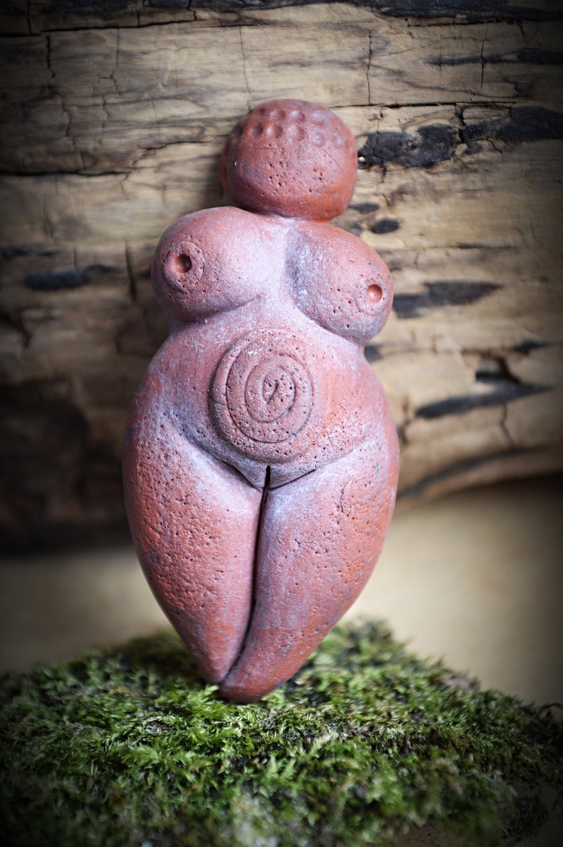 May include: A terracotta figurine of a female figure with a circular design on the abdomen. The figurine is standing on a bed of green moss.