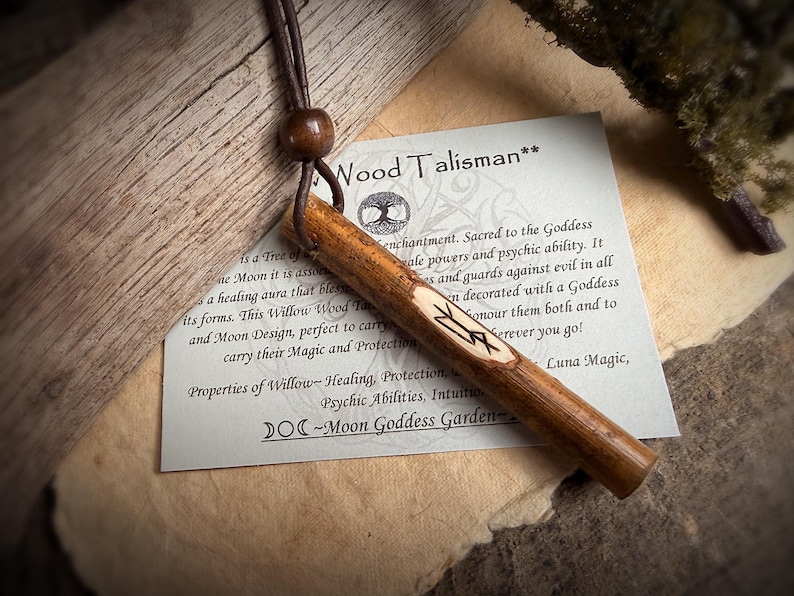 K&ouml;nnte beinhalten: Ein h&ouml;lzernes Talisman-Anh&auml;nger mit einer Lederschnur und einer Holzperle. Der Talisman ist zylindrisch, mit geschnitzten Symbolen und Text auf einem Papieranh&auml;nger mit der Aufschrift "Wood Talisman". Der Anh&auml;nger ist braun und beige.