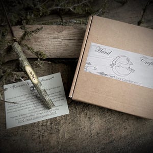 May include: A handcrafted wooden wand with a carved star design, accompanied by a card and a cardboard box with a label that reads "Hand Crafted" and "Moon Goddess Garden.com". The wand is made of natural wood.