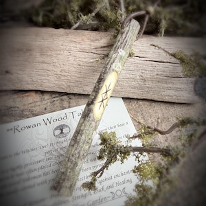 May include: A Rowan Wood Talisman, a wooden stick with carved runes, rests on a wooden surface with moss and a printed card. The card reads "Rowan Wood Talisman" and describes the tree's protective properties. The talisman is a natural, rustic item.