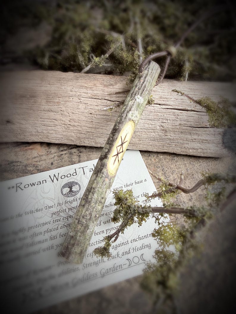 May include: A Rowan Wood Talisman, a wooden stick with carved runes, rests on a wooden surface with moss and a printed card. The card reads "Rowan Wood Talisman" and describes the tree's protective properties. The talisman is a natural, rustic item.