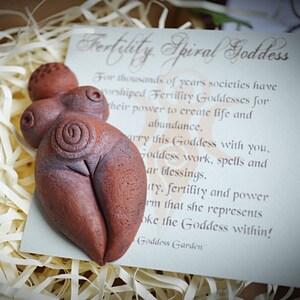 May include: A terracotta fertility goddess figurine with a spiral design on the abdomen. The figurine is sitting on a white card with the text "Fertility Spiral Goddess" and a description of the goddess.