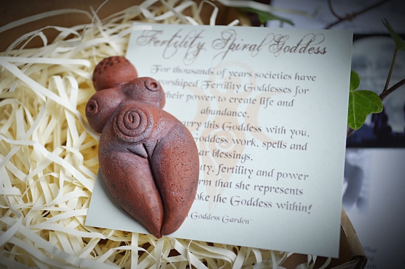 May include: A terracotta fertility goddess figurine with a spiral design on the abdomen. The figurine is sitting on a white card with the text "Fertility Spiral Goddess" and a description of the goddess.