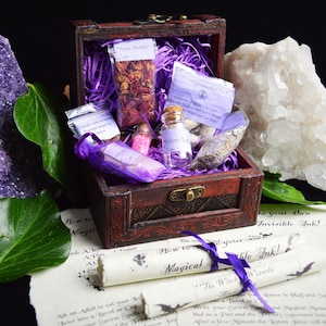 Op de afbeelding: Een houten schatkist gevuld met paarse en witte voorwerpen, waaronder gedroogde kruiden, een klein flesje glitter en een klein flesje vloeistof. De kist is open en staat op een wit oppervlak met een rol die zegt "Hoe maak je je eigen onzichtbare inkt!"