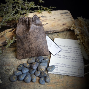 May include: A set of rune stones with gold symbols, a brown leather pouch, and informational cards. The stones are gray and arranged on a wooden surface. The text "Moon Goddess Garden" is at the bottom.