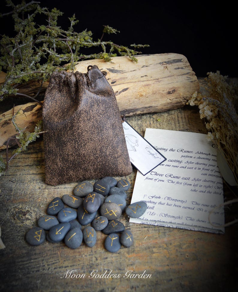 May include: A set of rune stones with gold symbols, a brown leather pouch, and informational cards. The stones are gray and arranged on a wooden surface. The text "Moon Goddess Garden" is at the bottom.