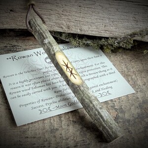 May include: A Rowan wood talisman with a brown leather thong. The wooden talisman has a light-colored oval with runes carved into it. A card with text about the talisman is next to it.