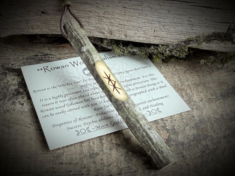 May include: A Rowan wood talisman with a brown leather thong. The wooden talisman has a light-colored oval with runes carved into it. A card with text about the talisman is next to it.