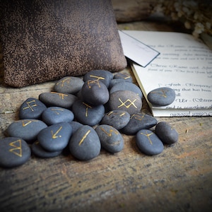 May include: A collection of gray, rune-inscribed stones with gold lettering. A brown pouch and a page of handwritten text are in the background. The stones are arranged on a wooden surface, suggesting a mystical or divination theme.