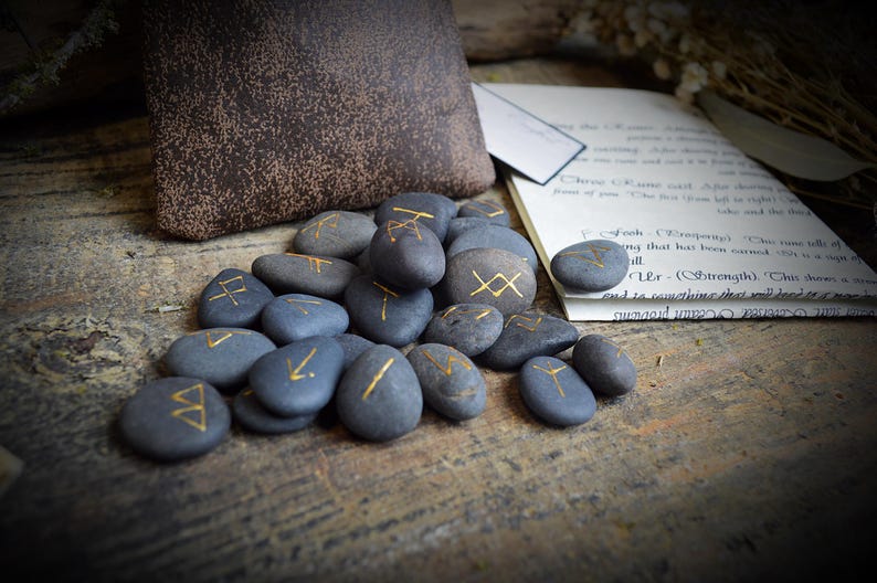 May include: A collection of gray, rune-inscribed stones with gold lettering. A brown pouch and a page of handwritten text are in the background. The stones are arranged on a wooden surface, suggesting a mystical or divination theme.