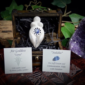 May include: White ceramic figurine of a goddess with a blue sodalite crystal in her belly. The figurine is in a wooden box with black shredded paper. The box is sitting on a book with a purple amethyst crystal next to it. The text on the card reads "Air Goddess *Luna* with Sodalite Crystal -by Moon Goddess Garden- *Sodalite* For Self Esteem and Communication. Helps with Intuition"