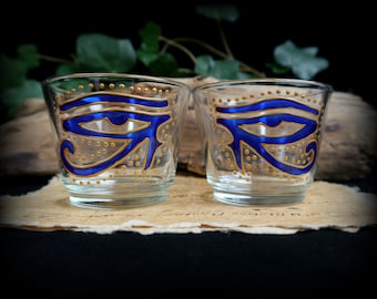 May include: Two clear glass candle holders with a blue and gold painted Eye of Horus design.