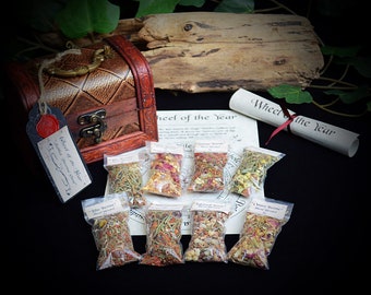 May include: A wooden chest with a brass latch and a tag that reads "Wheel of the Year" sits on a black surface.  Eight small bags of dried herbs and flowers are arranged in front of the chest.  The bags are labeled with the names of the eight sabbats of the Wheel of the Year.