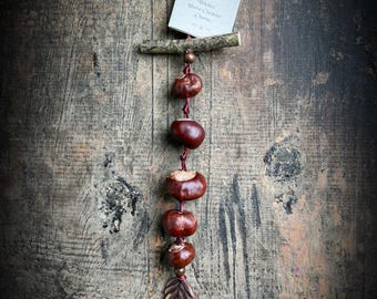 May include: A hanging ornament featuring five polished brown horse chestnuts strung on a dark red ribbon. The ornament includes a small horizontal wooden twig, a square tag with the text Witches Horse Chestnut Charm, and a metal leaf charm.