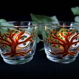 Könnte beinhalten: Zwei klare Glas-Kerzenhalter mit einem gemalten Design eines Baumes mit grünen Blättern und einer goldenen Umrandung. Der Baum ist von einem weißen Kreis mit goldenen Punkten umgeben. Die Kerzenhalter sind mit Teelichtern beleuchtet.