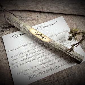 May include: A Rowan wood talisman with a leather thong, featuring a carved rune. The talisman is accompanied by a card with text about Rowan's protective properties and the phrase "Moon Goddess Garden."