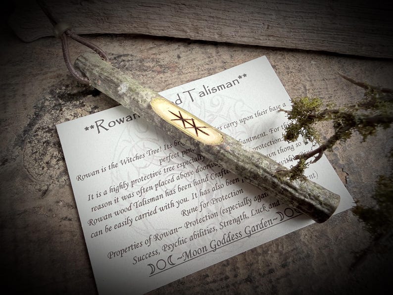 May include: A Rowan wood talisman with a leather thong, featuring a carved rune. The talisman is accompanied by a card with text about Rowan's protective properties and the phrase "Moon Goddess Garden."