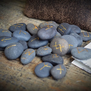 May include: A collection of gray stones with gold-colored runic symbols. The stones are arranged on a wooden surface, with a brown pouch and a piece of paper in the background. The runes are painted in a gold color.