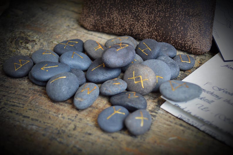 May include: A collection of gray stones with gold-colored runic symbols. The stones are arranged on a wooden surface, with a brown pouch and a piece of paper in the background. The runes are painted in a gold color.
