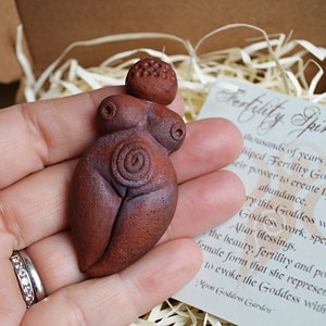 May include: A small, hand-crafted terracotta figurine of a fertility goddess. The figurine is brown and has a spiral design on its belly. The figurine is being held in a hand. The text on the card reads "Fertility Spiral Goddess".