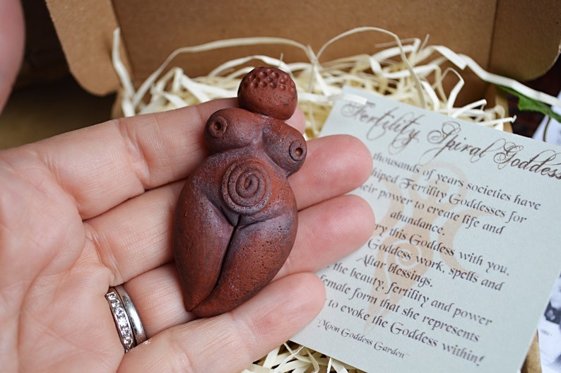 May include: A small, hand-crafted terracotta figurine of a fertility goddess. The figurine is brown and has a spiral design on its belly. The figurine is being held in a hand. The text on the card reads "Fertility Spiral Goddess".