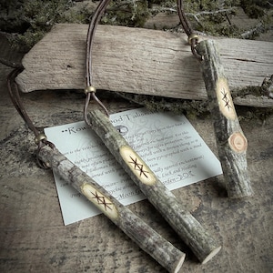 May include: Three wooden pendants with leather cords. Each pendant is a light brown color with a carved design and the text "Rowan Wood Talisman". The pendants are displayed on a wooden surface with foliage.