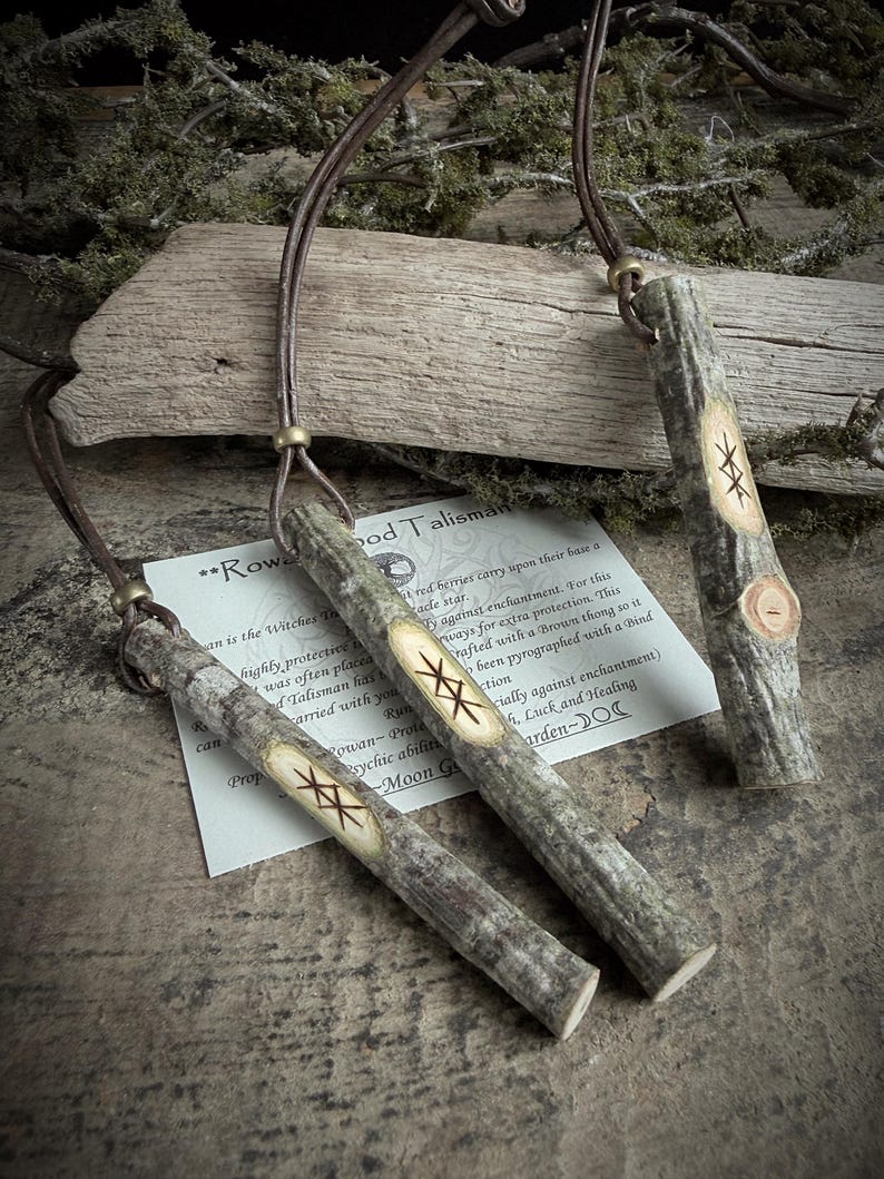 May include: Three wooden pendants with leather cords. Each pendant is a light brown color with a carved design and the text "Rowan Wood Talisman". The pendants are displayed on a wooden surface with foliage.