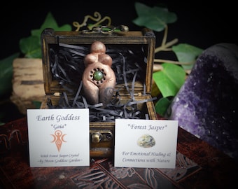 May include: A copper-colored Earth Goddess figurine with a green forest jasper crystal in its belly. The figurine is in a wooden box with black shredded paper. The box is sitting on a wooden surface with a purple amethyst crystal in the background.