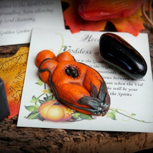 May include: Orange and black clay sculpture of a goddess figure with a black stone in the centre. The sculpture is on a white card with text that reads "Hecate Goddess of the Night".