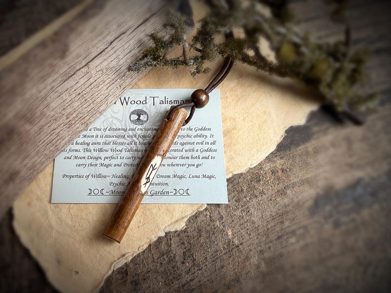 K&ouml;nnte beinhalten: Ein h&ouml;lzernes Talisman mit einer Lederschnur und einer Holzperle. Das Talisman ist mit einem Symbol graviert und liegt auf einem Papier mit Text &uuml;ber seine Eigenschaften. Der Hintergrund ist eine rustikale Holzoberfl&auml;che.