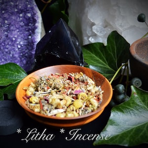 May include: A wooden bowl filled with a blend of dried herbs and flowers, labeled "Litha* Incense". The bowl is surrounded by green leaves, a purple amethyst crystal, a black obsidian crystal, and a small brown mortar.