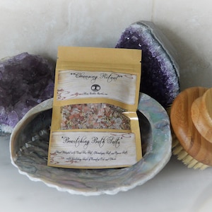 May include: A small brown paper bag with a label that reads "Cleansing Ritual" and "Bewitching Bath Salts". The bag is filled with pink bath salts and is sitting on a white surface next to a large amethyst crystal and a wooden bath brush.