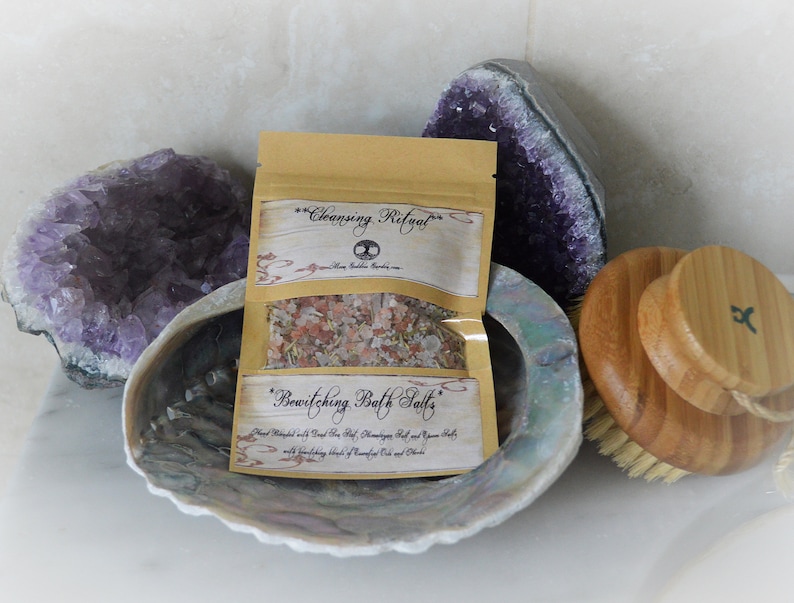 May include: A small brown paper bag with a label that reads "Cleansing Ritual" and "Bewitching Bath Salts". The bag is filled with pink bath salts and is sitting on a white surface next to a large amethyst crystal and a wooden bath brush.