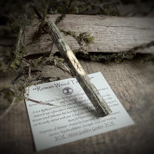 May include: A Rowan wood talisman with a carved rune, resting on a card with text about its protective properties. The talisman is light brown with a carved symbol. The card is white with black text.
