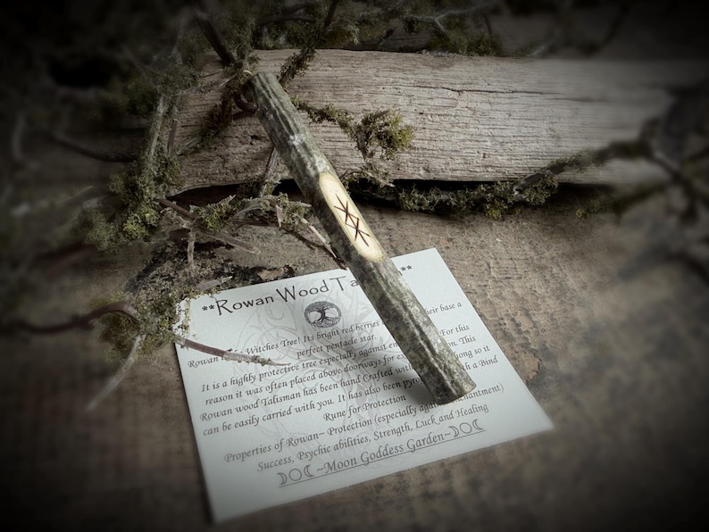 May include: A Rowan wood talisman with a carved rune, resting on a card with text about its protective properties. The talisman is light brown with a carved symbol. The card is white with black text.