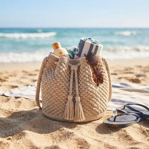 May include: A beige crocheted bucket bag with a drawstring closure and two tassels. Inside the bag are a white and blue striped towel and a bottle of sunscreen. A pair of brown tortoiseshell sunglasses rests on the bag, and blue flip-flops sit nearby.
