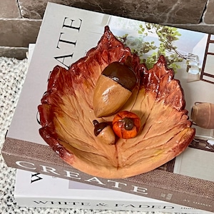 Puede incluir: Un plato decorativo de cerámica con forma de hoja de otoño grande con un degradado de colores naranja, rojo y marrón. Dentro del plato hay tres objetos esculpidos: una bellota marrón grande, una bellota marrón más pequeña y una calabaza redonda.