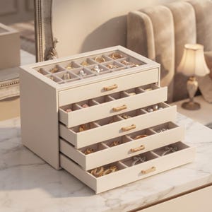 May include: A cream-colored jewelry box with a glass top lid and four pull-out drawers. Each drawer and the top compartment feature multiple square dividers for organizing accessories. The box has gold-toned metal handles on each drawer front.