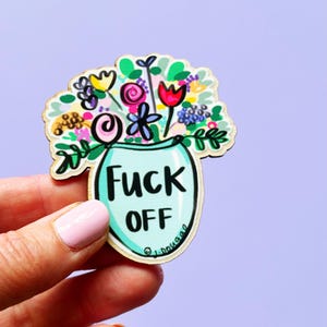 May include: A wooden pin featuring a colourful illustration of a bouquet of flowers in a light blue vase. The vase has the text FUCK OFF printed in bold black letters. The flowers are depicted in shades of red, yellow, pink, purple, and green.