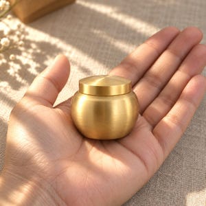 May include: A small, round, brushed brass container with a matching lid. The vessel features a smooth, curved body and a flat, circular top, showcasing a metallic gold finish with a subtle, matte texture throughout the entire exterior surface.