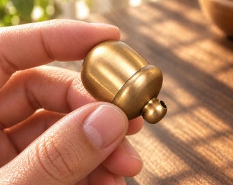 May include: A small, smooth, acorn-shaped brass container with a polished metallic finish. The object features a rounded top section and a slightly smaller base with a small circular opening at the bottom, designed as a compact, solid metal charm.