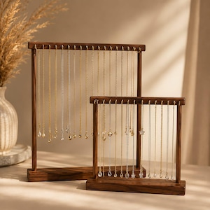 May include: Two rectangular dark wood jewelry stands with horizontal bars featuring multiple gold-toned metal hooks. Numerous delicate gold and silver chains with small clear crystal or metallic pendants hang vertically from the hooks on both stands.